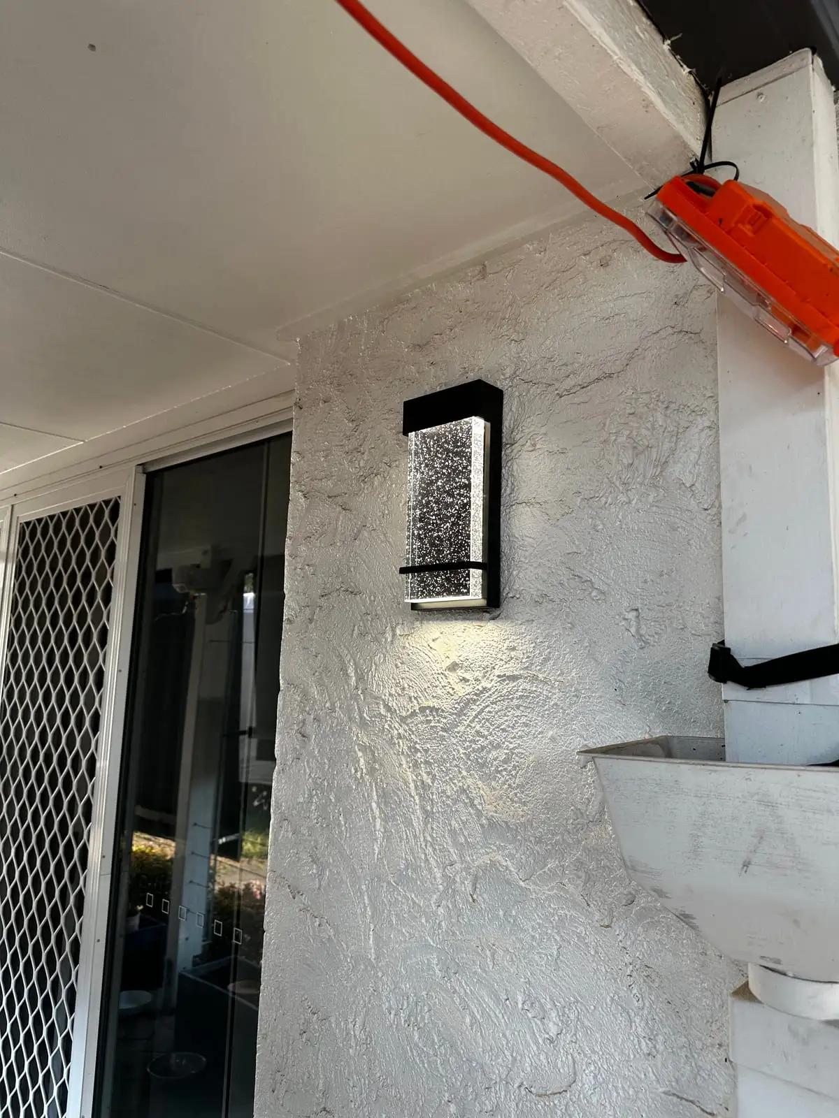 A rectangular black LED wall light lit on a textured white rendered patio wall beside a sliding door
