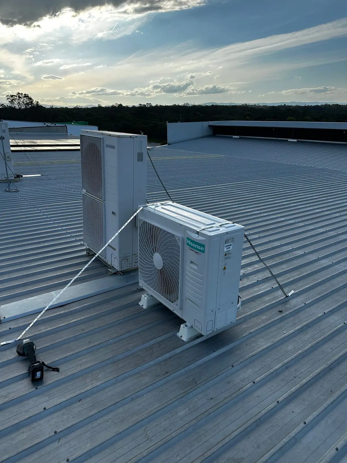 Two white air conditioning condensers mounted and strapped down on a large commercial metal roof at dusk