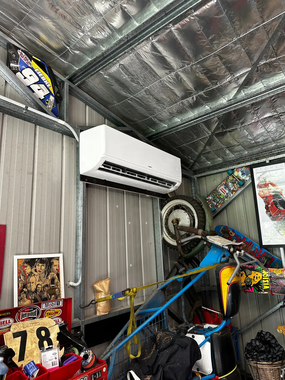 A white split system head unit mounted high in the corner of a colourbond shed above a workbench and a dirt bike