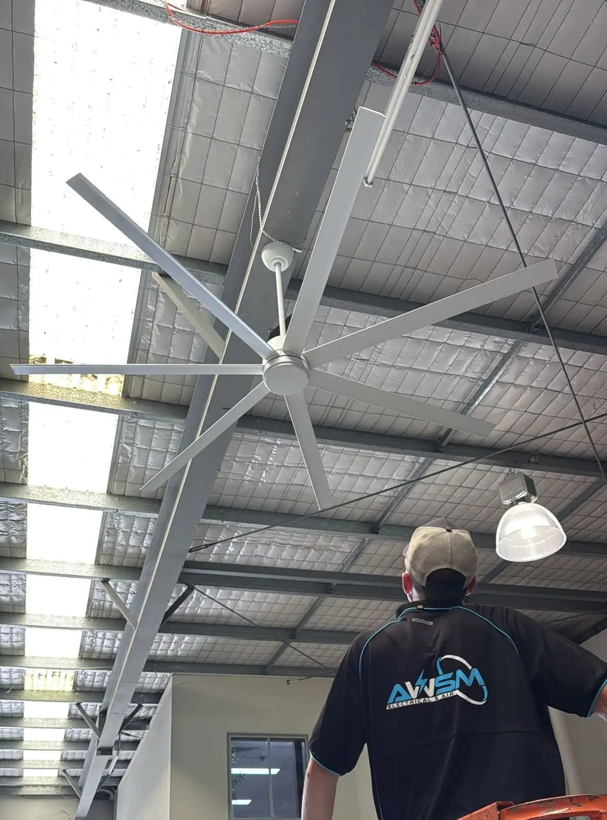 A large six-blade high-volume low-speed fan installed under a warehouse roof, an AWSM electrician on a scissor lift below it