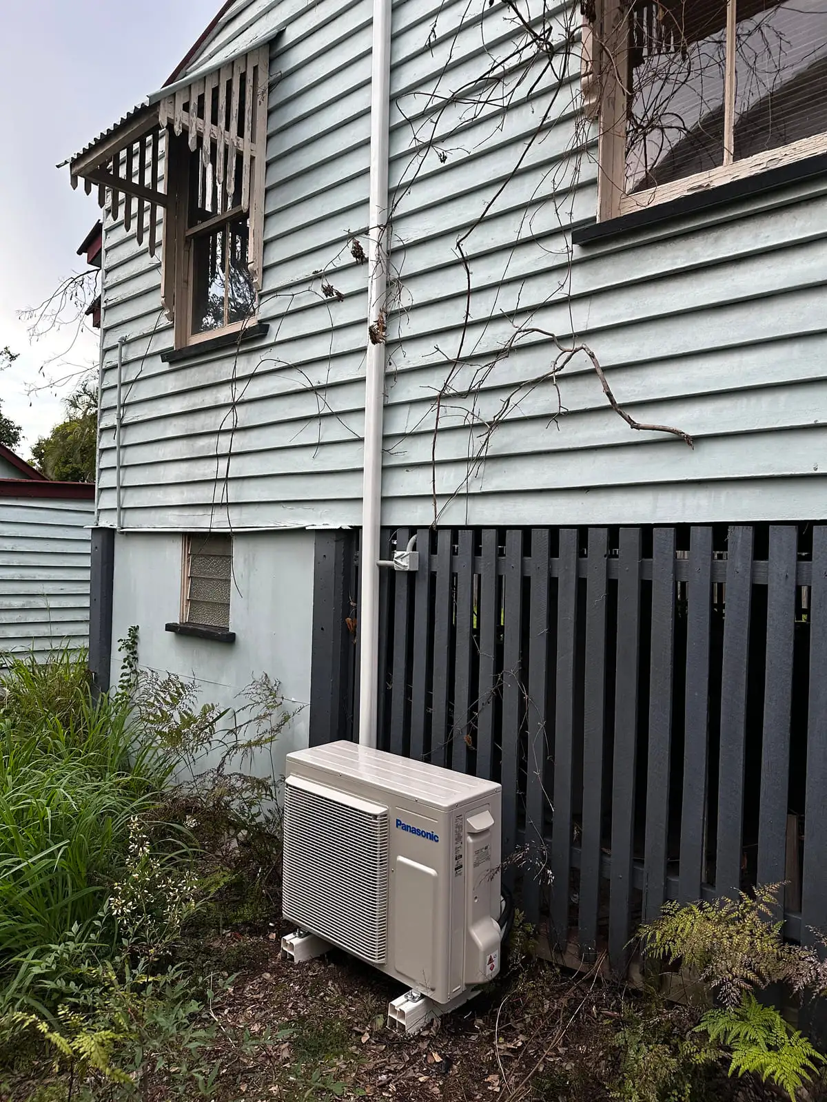 A Panasonic outdoor air conditioning unit on feet in a garden bed beside a pale blue weatherboard Queenslander, pipework run up the wall in white ducting