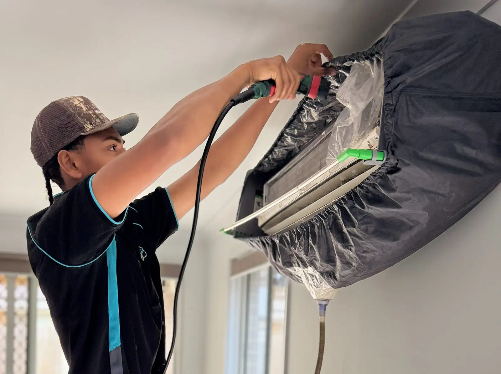 An AWSM tech washing a split system's coil, the head wrapped in a cleaning bag with a drain hose running down the wall