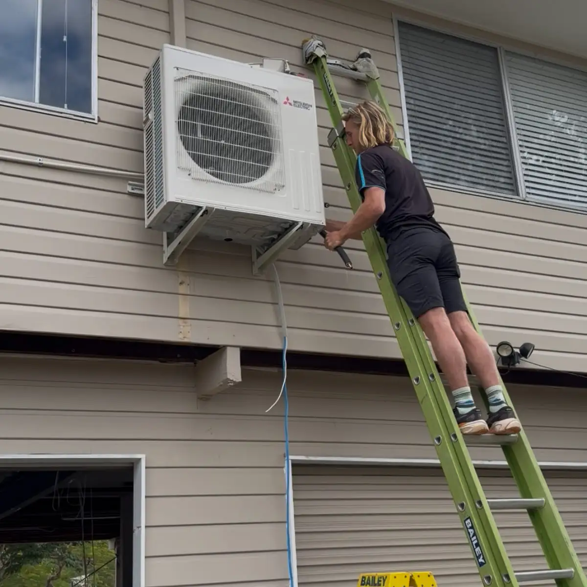 An AWSM installer up a ladder bolting a Mitsubishi Electric outdoor unit to a weatherboard wall