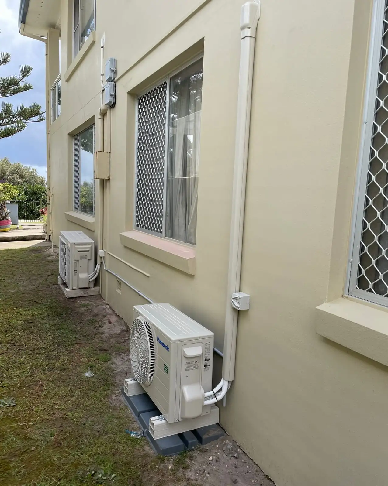 Two Panasonic outdoor air conditioning units on brackets beside a rendered wall, pipework run in tidy white ducting