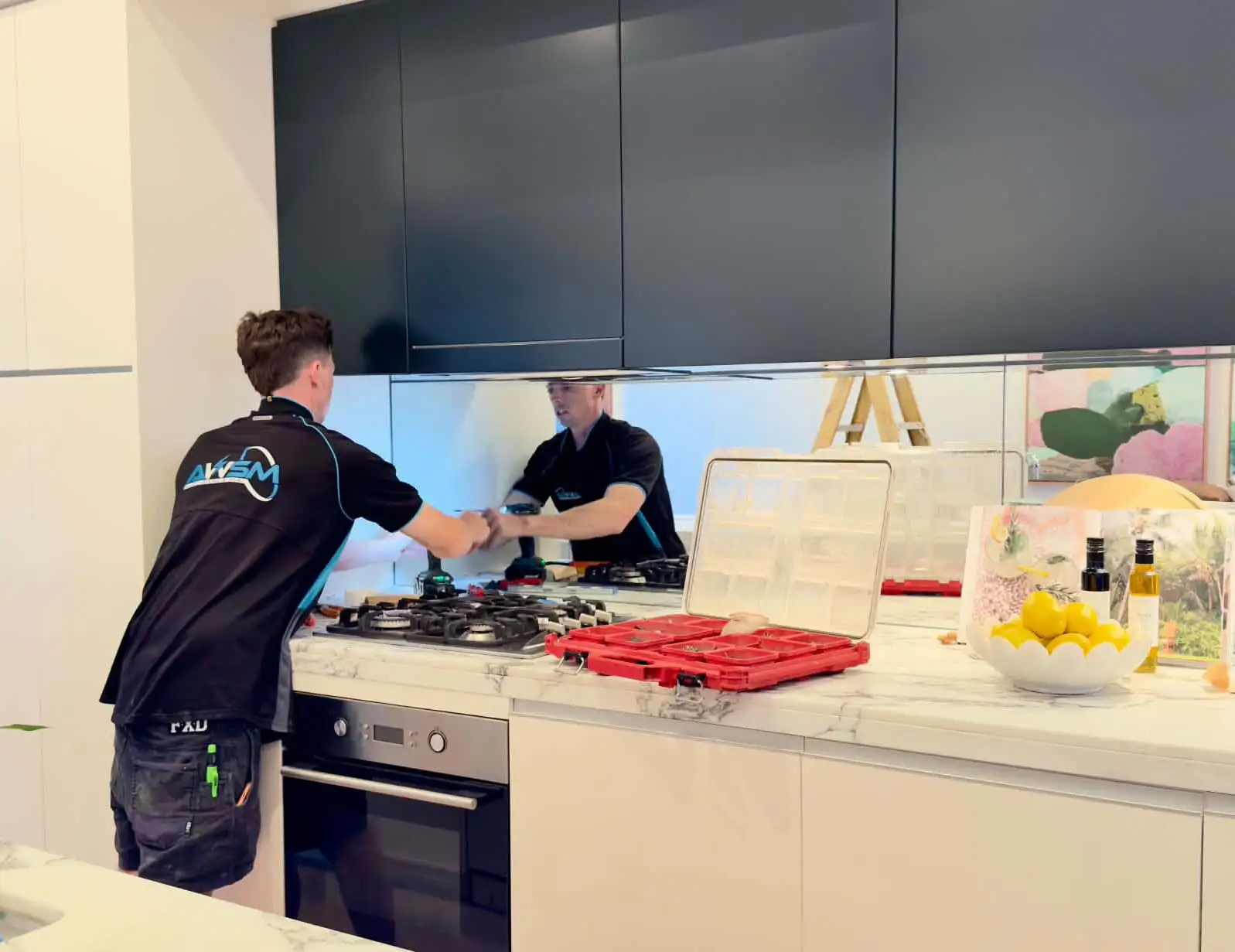 An AWSM electrician working at a mirrored kitchen splashback above the cooktop, drill in hand, a red parts case open on the benchtop
