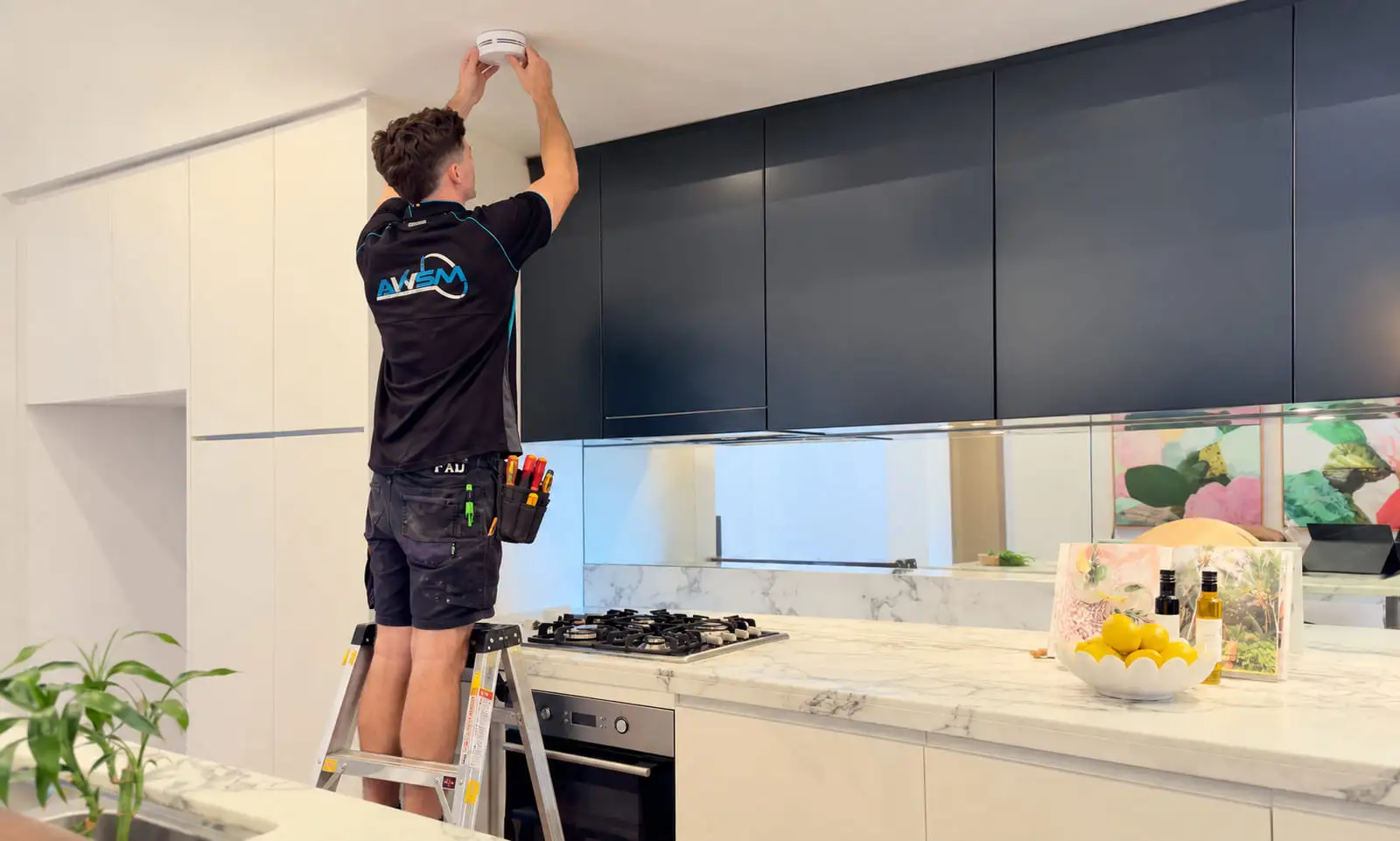 An AWSM electrician on a step ladder fitting a smoke alarm to the ceiling of a kitchen