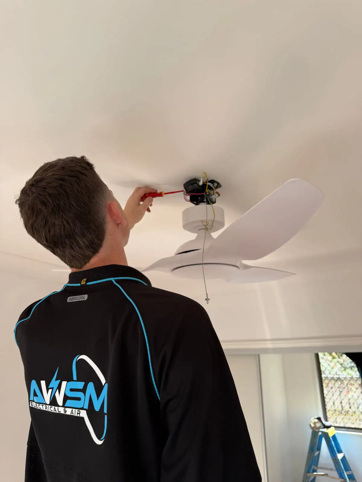 An AWSM electrician in a branded shirt wiring the fan at the ceiling, stepladder beside him